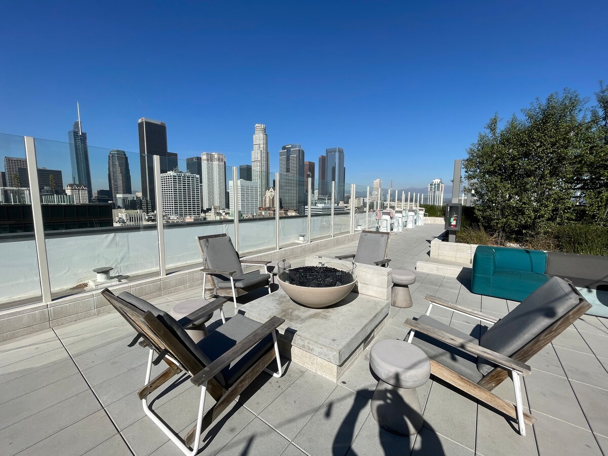 A rooftop area provides stunning city skyline views, featuring several lounge chairs arranged around a circular fire pit. A small table is positioned nearby, while lush greenery is visible in one corner, enhancing the open-air space.