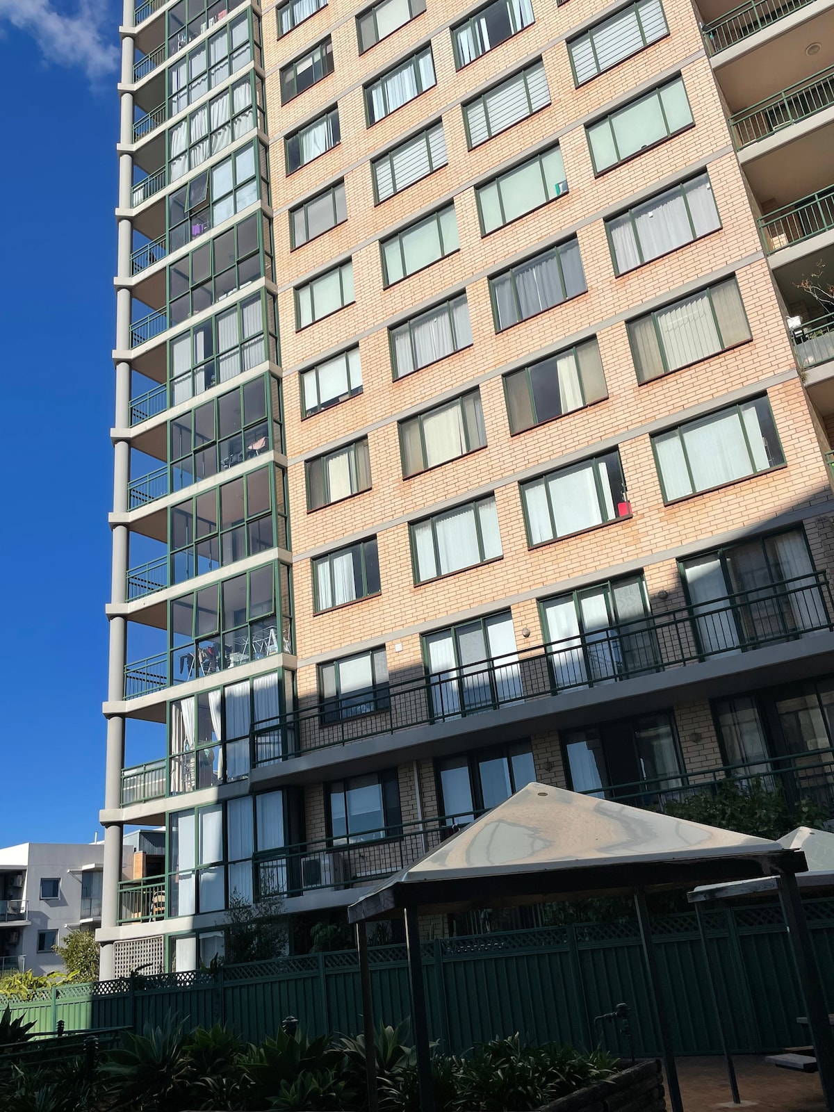 The multi-story apartment building is characterized by large glass windows and a combination of brick and modern design. A shaded outdoor seating area is situated in the foreground, framed by well-maintained greenery.