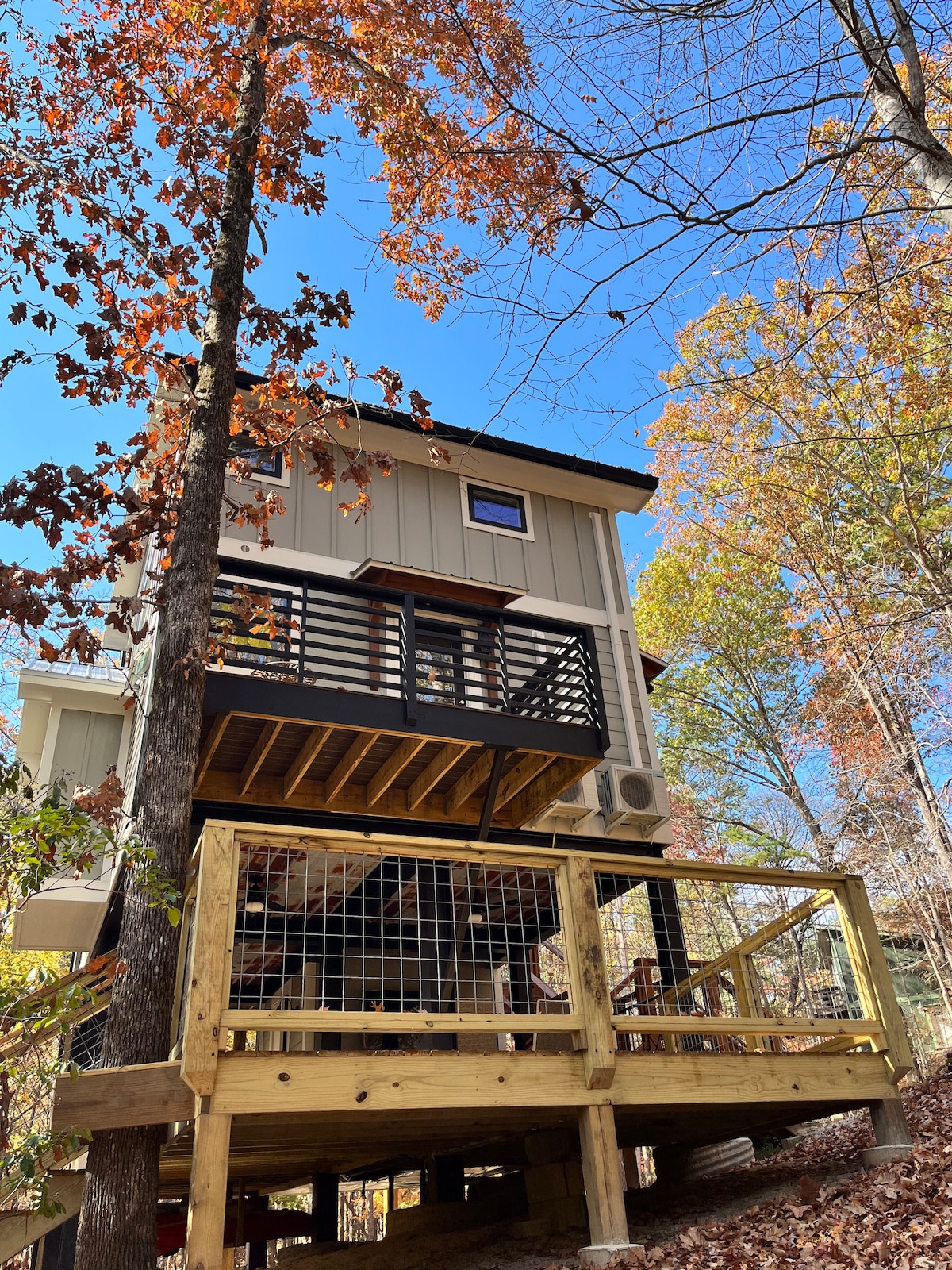 A modern two-story home stands nestled among vibrant autumn trees, showcasing a spacious deck supported by wooden beams. The exterior features a blend of earthy tones, with large windows allowing natural light. A clear blue sky creates a serene backdrop.