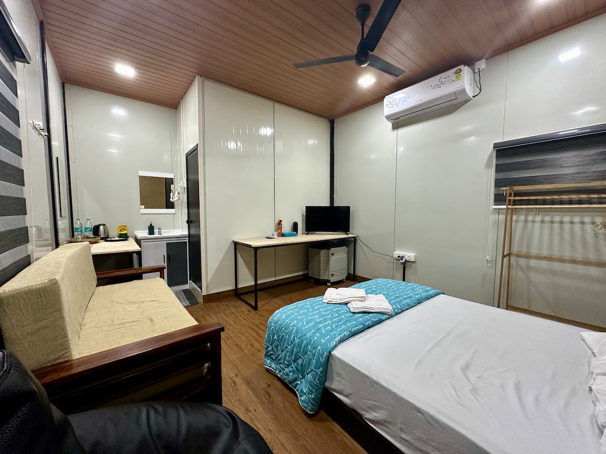 The room offers a spacious layout featuring a double bed with a light blue quilt. A small sofa sits against one wall, alongside a desk and chair. A flat-screen TV is visible on a stand, while a mini-fridge is positioned nearby.