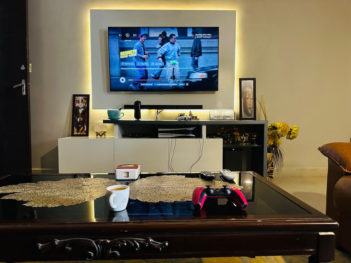 A modern living area features a mounted flat-screen TV displaying programming. A sleek console underneath holds multimedia devices and decor. A coffee table with an elegant design is centered in front, with a cup placed on it and a gaming controller positioned nearby.