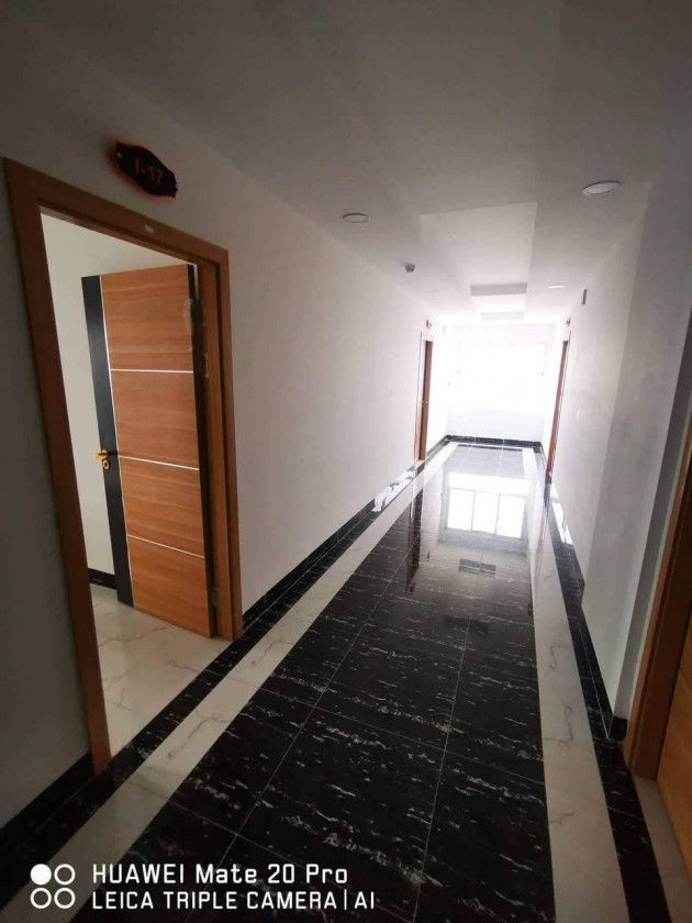 A long corridor is shown with glossy black and white tiled flooring reflecting soft overhead lighting. Doorways are visible on either side, leading to individual rooms. Natural light filters in from the end, creating a welcoming and open space.
