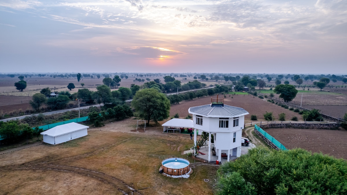 An aerial view captures the unique octagonal farmhouse surrounded by expansive fields. A small circular swimming pool is positioned nearby, reflecting the soft colors of the sunset. The scene is framed by lush trees and open land, creating a serene atmosphere.