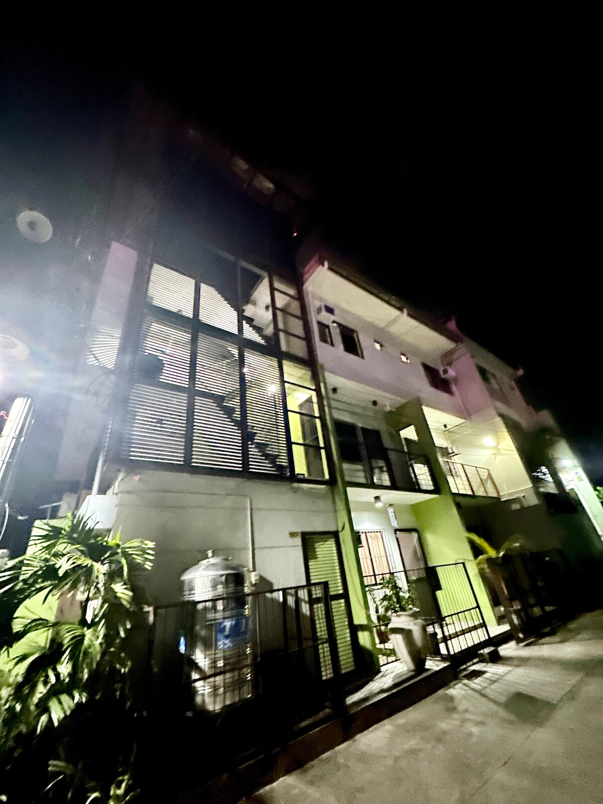 A multi-story building is viewed at night, showcasing a modern facade with large windows and an upper-level balcony. Soft lighting illuminates the entrance area, complemented by greenery located near the entrance. The exterior features a blend of light and dark tones.