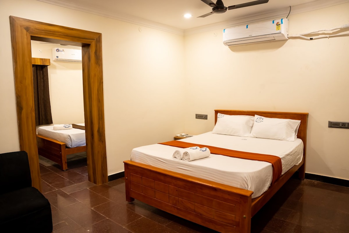 A well-lit bedroom features a spacious bed with stylish wooden framing, complemented by crisp white linens and a contrasting orange throw. A mirror reflects an additional bed in the background. An air conditioning unit is mounted on the wall, and a comfortable chair is positioned in the corner.