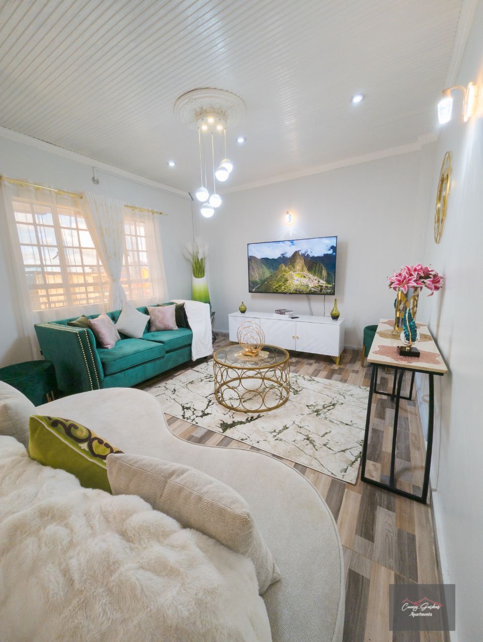 A bright living area features a teal sofa and a light-colored rug, complementing the neutral walls. A television is mounted on the wall, while a decorative table holds a vase of flowers. Natural light filters through the curtains, highlighting the cozy atmosphere.