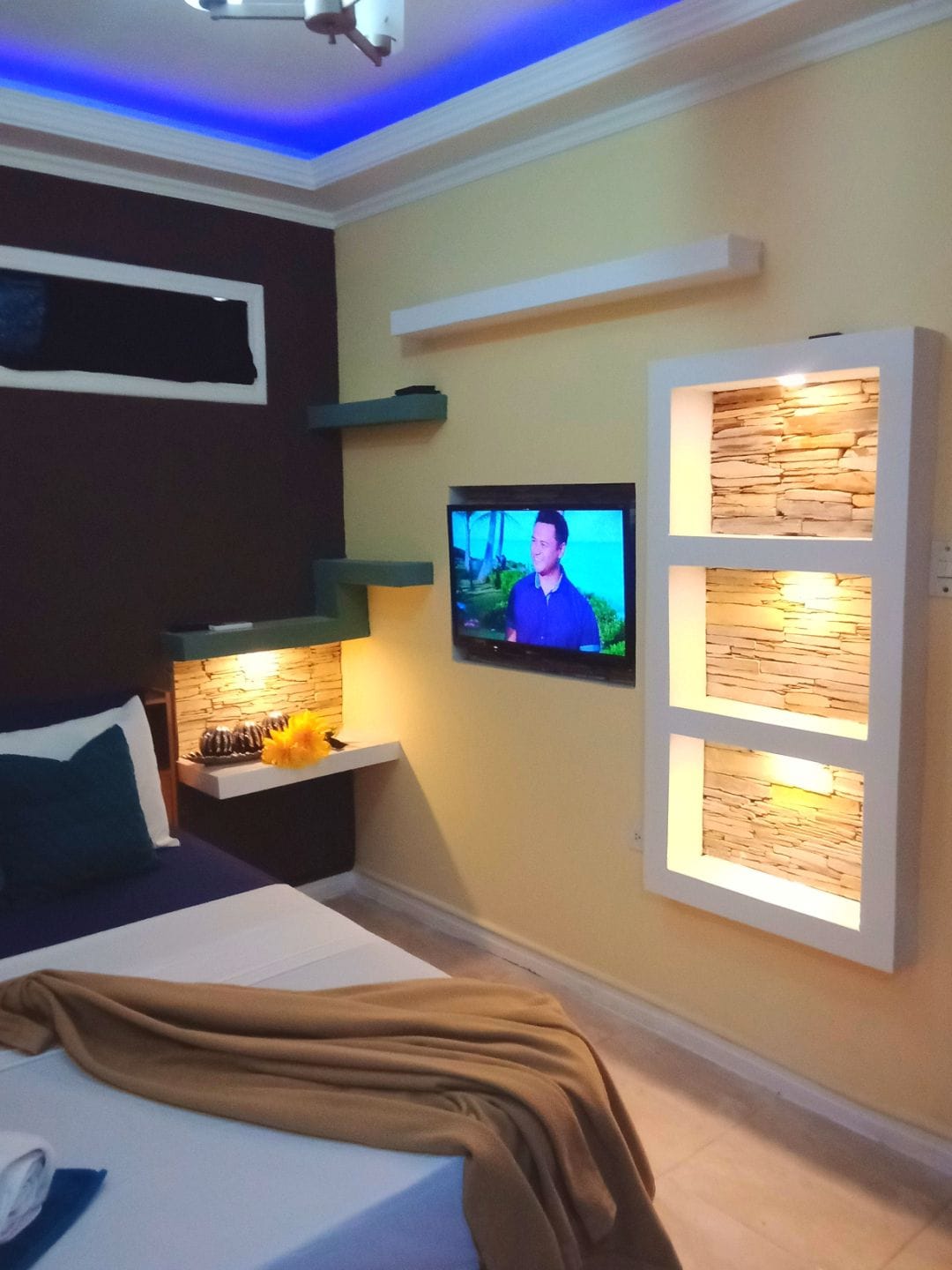 A cozy room is presented with a spacious bed featuring a neatly arranged blanket. A flat-screen TV is mounted on the wall, accompanied by stylish shelves displaying decorative elements. Soft lighting enhances the design, creating a warm and comfortable environment.