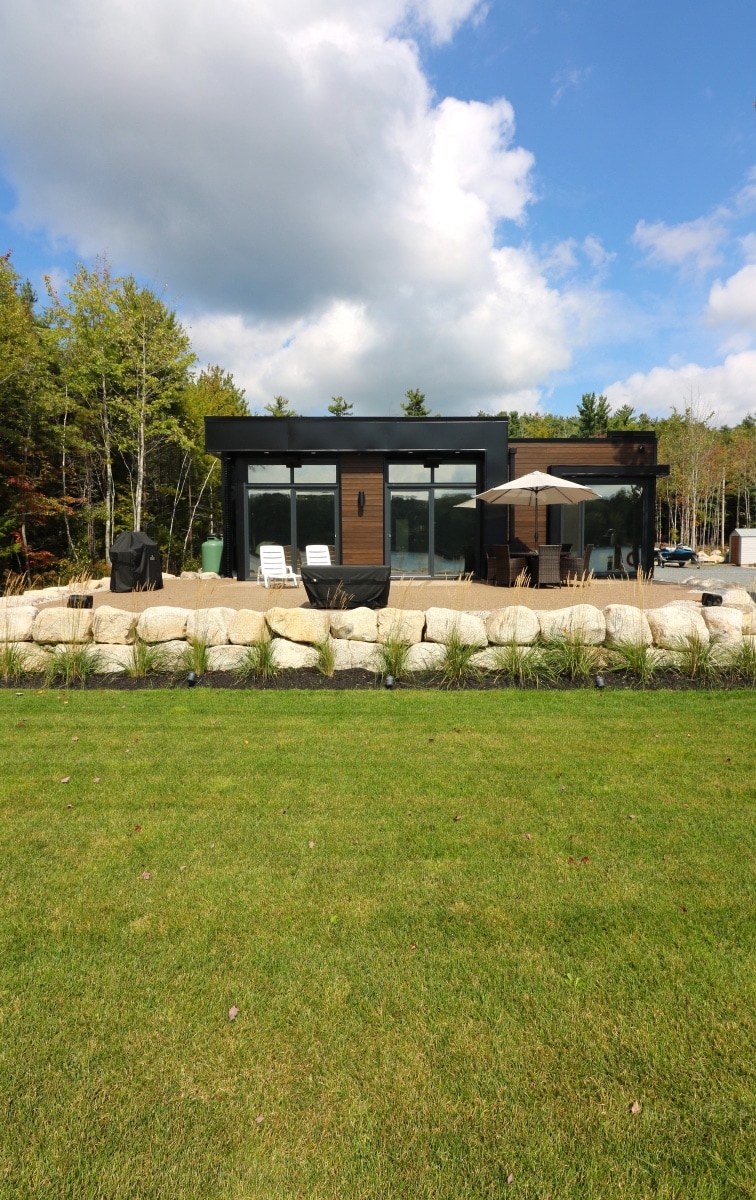 A modern structure is seen with large windows, allowing natural light to fill the interior. The surrounding area features well-maintained grass and decorative boulders, complemented by trees in the background under a partly cloudy sky. Outdoor furniture is positioned for relaxation.