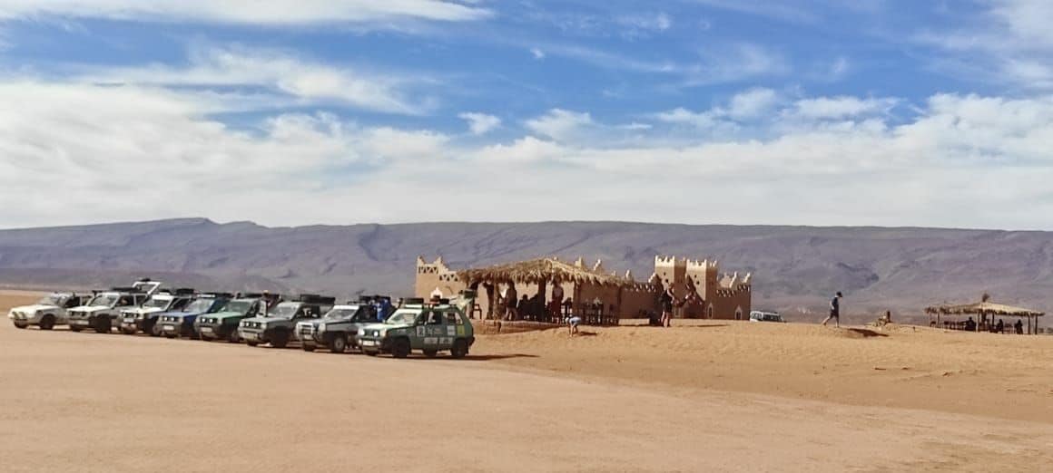 A row of vehicles is parked on sandy terrain, with a desert lodge visible in the background. The lodge features a thatched roof and walls made from earthy materials. Light clouds drift across a blue sky, while rugged mountains are seen in the distance.