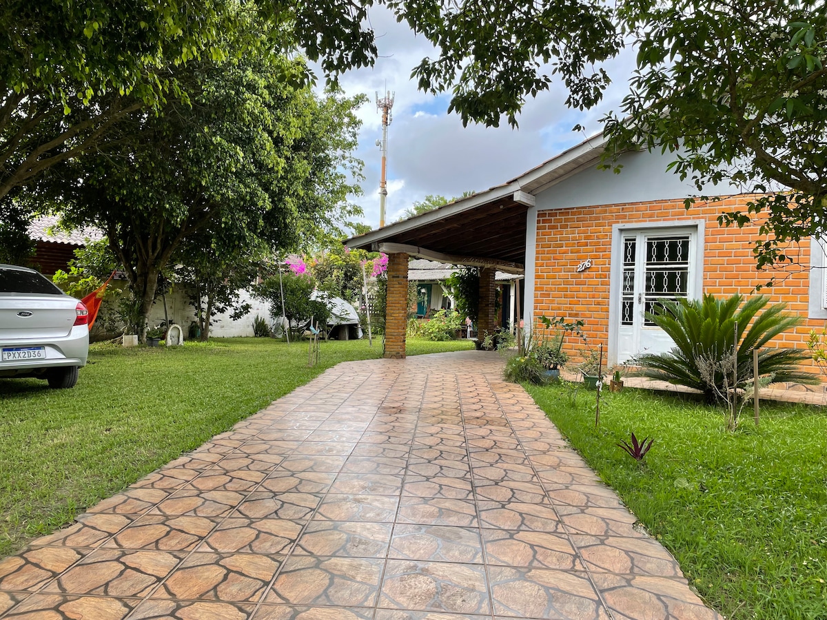 The entrance is framed by a well-maintained garden featuring lush green grass and trees. A tiled walkway leads to the house, which has a covered porch and a brick façade. Parking spaces are visible alongside the path.
