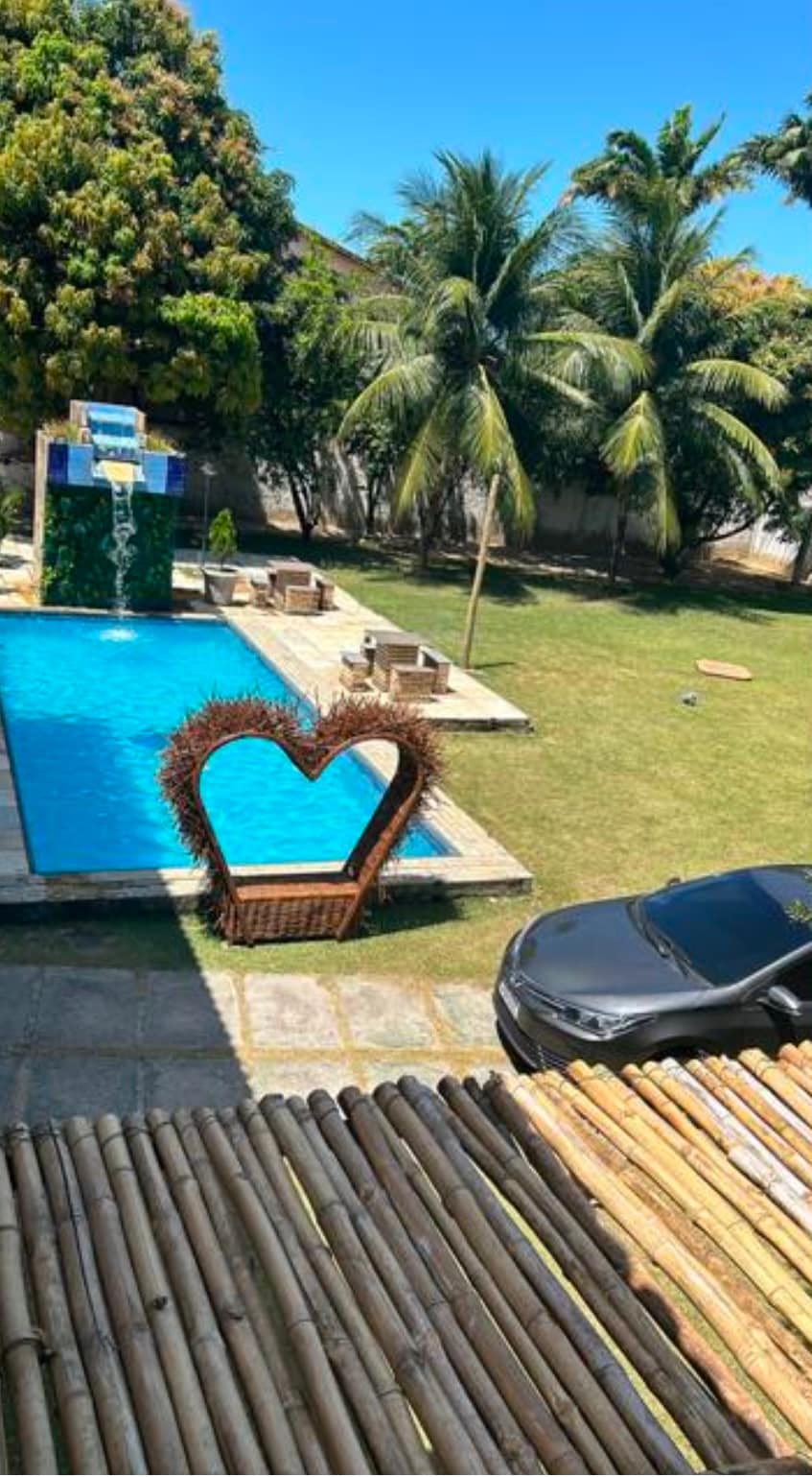 An outdoor area features a clear blue pool surrounded by sun loungers and a large grassy space. A heart-shaped decorative feature stands near the pool, while tropical palm trees provide shade. A vehicle is parked nearby, with a bamboo structure in the foreground.