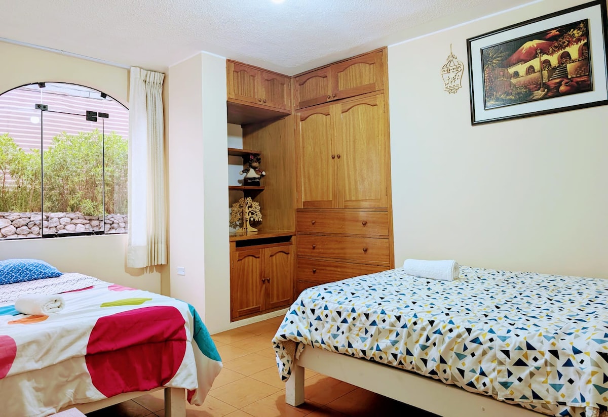 A bright bedroom features two beds with colorful bedding, one adorned with polka dots and the other with a geometric pattern. Natural light enters through a large arched window, framed by light curtains, while wooden cabinetry provides practical storage along one wall.