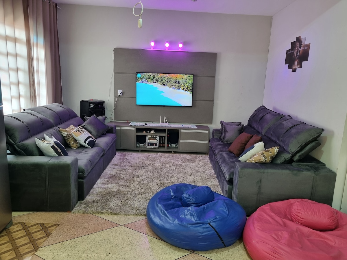 The living room features two spacious sofas facing a wall-mounted television. A soft area rug covers the floor, and comfortable bean bags are positioned nearby in vibrant blue and pink. Natural light filters through the curtains, adding warmth to the space.