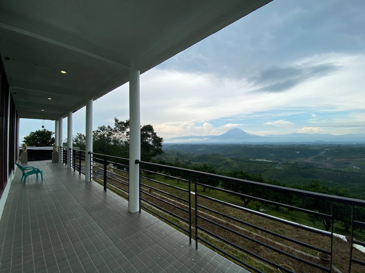 A spacious terrace is presented, featuring a tiled floor and a railing that overlooks expansive green landscapes and distant mountains. Comfortable seating is arranged, inviting relaxation while enjoying the sweeping views. The sky displays a blend of soft clouds and sunlight.