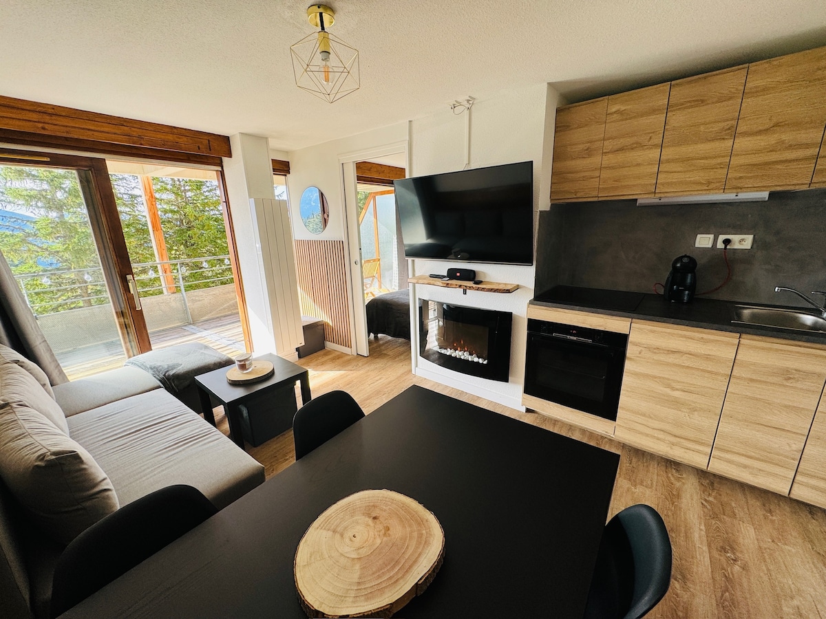 The open-concept living area features a comfortable couch and a dining table, complemented by a wall-mounted television. A cozy electric fireplace offers warmth. Large windows provide unobstructed views of the surrounding landscape, enhancing the bright and modern aesthetic of the space.