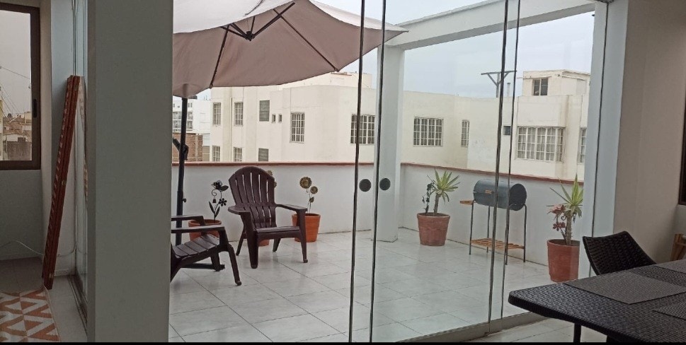 A spacious terrace is revealed, showcasing a small grill and multiple planters with greenery. Two comfortable chairs are positioned nearby under a large umbrella. Large glass doors invite natural light into the area, creating a bright and airy feel.