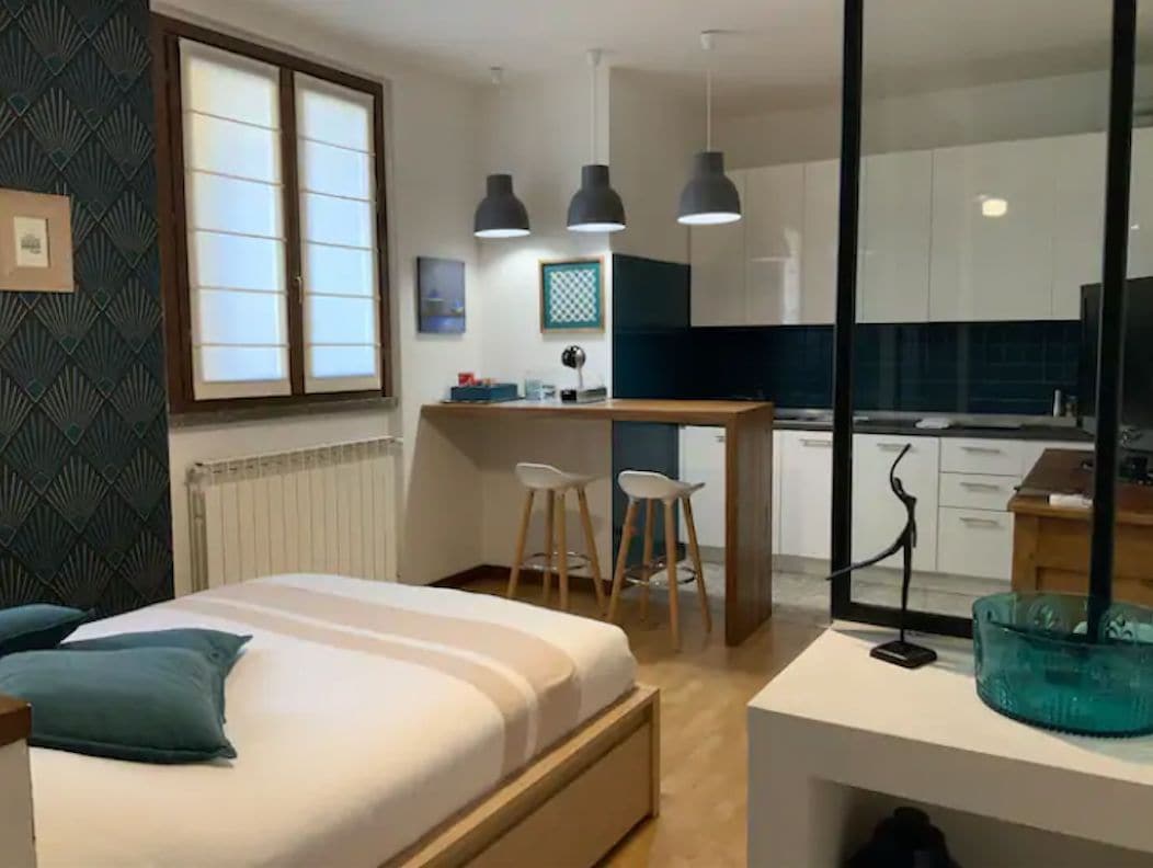 A spacious living area features a sleeping area with a simple bed and two coordinating pillows. A dining counter with two bar stools is visible, alongside a modern kitchen area with white cabinetry and clean blue accents. Large windows allow natural light to fill the space.