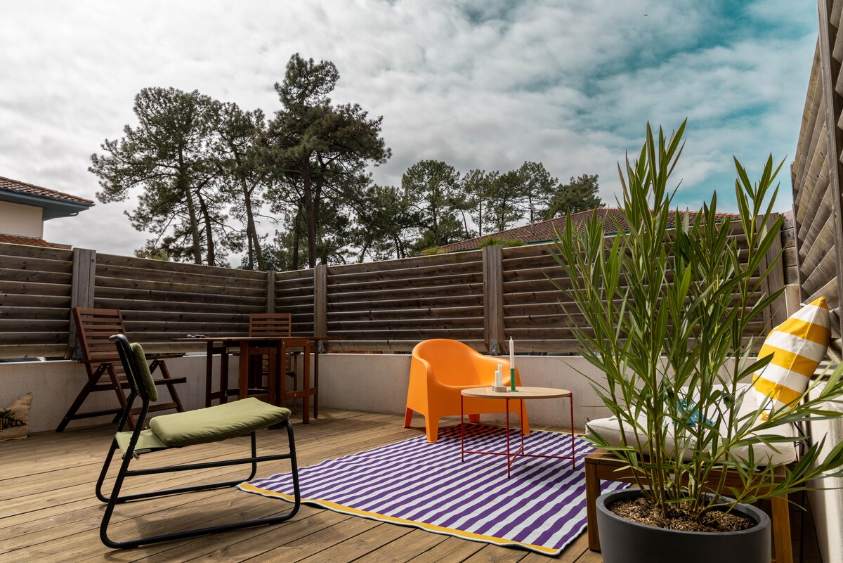 A spacious wooden terrace features a striped rug and a variety of seating options, including a green folding chair, an orange armchair, and a wooden table. Potted plants add a touch of greenery, while a wooden fence provides privacy against the backdrop of trees.