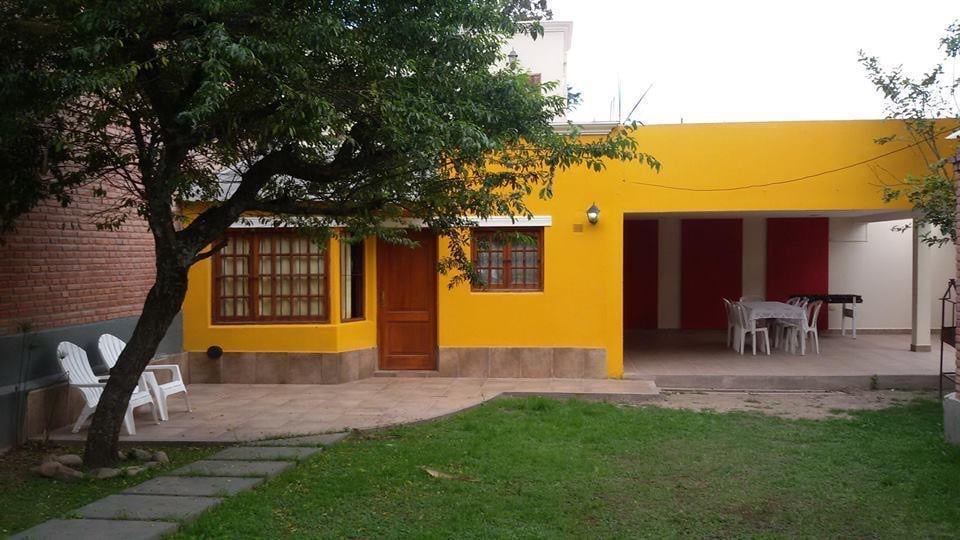 A vibrant yellow exterior of the house is framed by a lush green lawn and a large tree. The entrance features wooden doors and multiple windows, allowing natural light into the space. A shaded patio area includes a white table and chairs, ideal for outdoor gatherings.