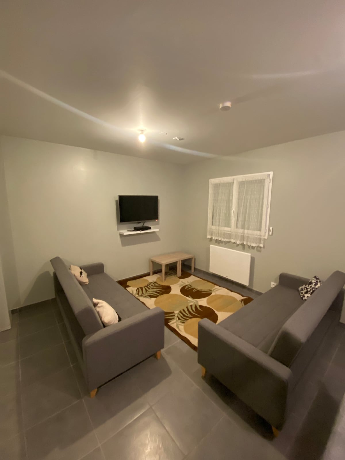A cozy living area features two gray sofas positioned around a low wooden table. A flat-screen television is mounted on the wall opposite. Light gray walls complement the tiled floor, while a patterned rug adds texture underfoot. A window with sheer curtains allows natural light to filter in.