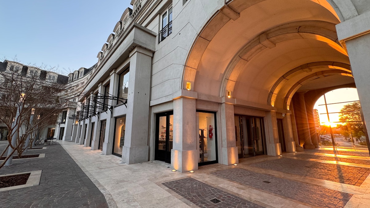 An exterior view of a modern building showcases a covered walkway with arched ceilings. Large glass doors are visible, providing glimpses into the interior. Soft golden light from the setting sun enhances the atmosphere, and a paved path leads through the surrounding landscaped area.