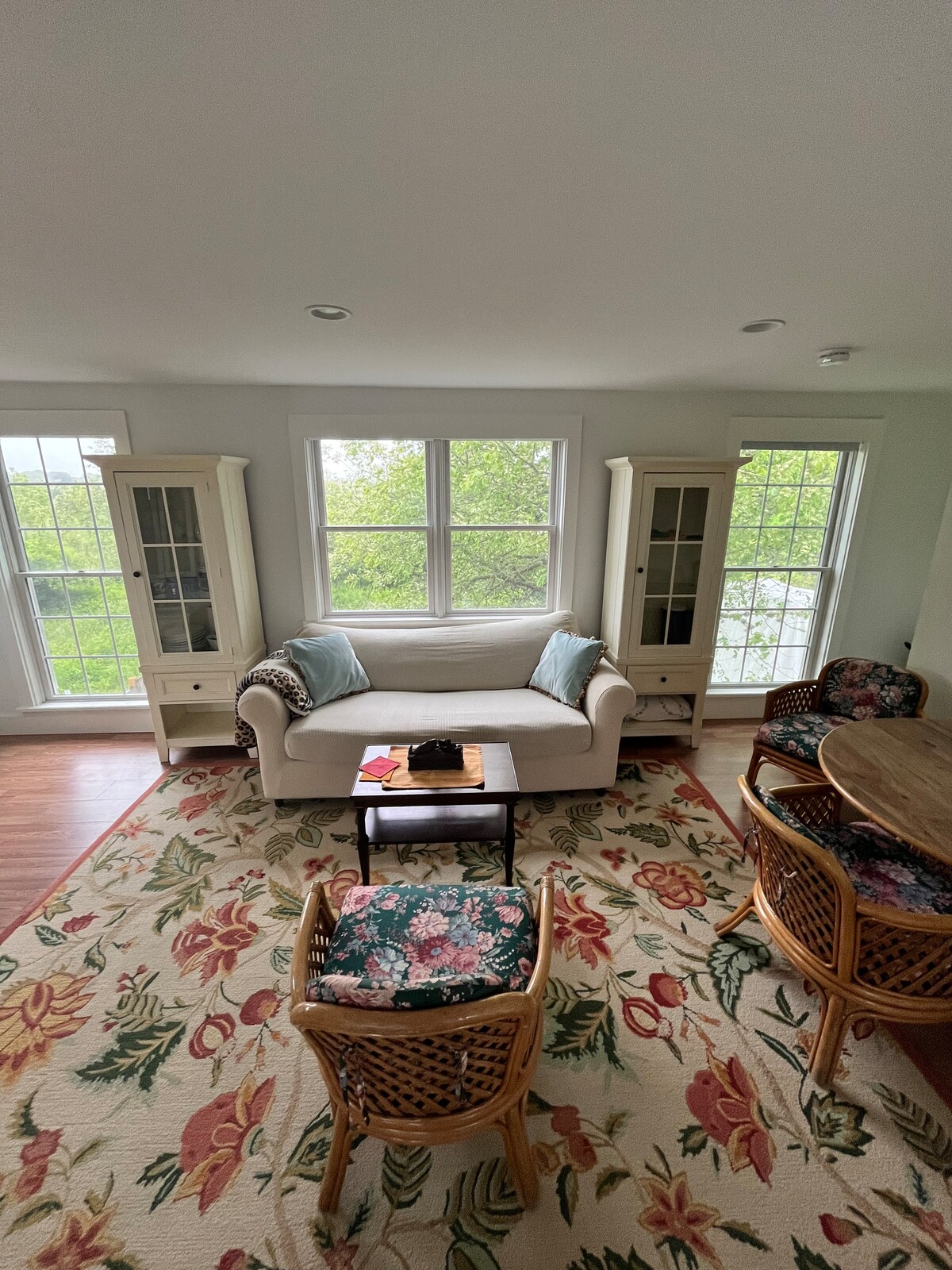 A light-filled living area includes a white couch adorned with colorful throw pillows, surrounded by two wicker chairs. A floral area rug adds warmth to the space. Two large windows provide a view of the outside greenery, enhancing the room's bright ambiance.