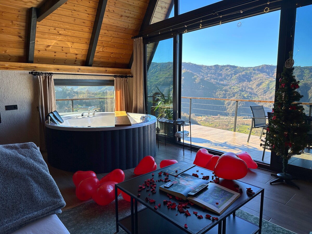 A spacious interior features a hydromassage jacuzzi alongside large glass doors that provide stunning views of the surrounding mountains. A coffee table is adorned with scattered petals, and a few red balloons are placed on the floor. A cozy bed is visible in the foreground.