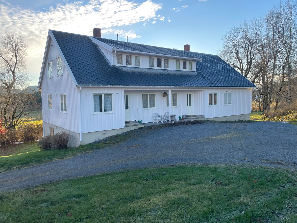 A large white house with a dark slate roof is set on a gently sloping green lawn. The structure features multiple windows that allow for natural light, and a gravel driveway leads to the entrance. Trees are visible in the background.