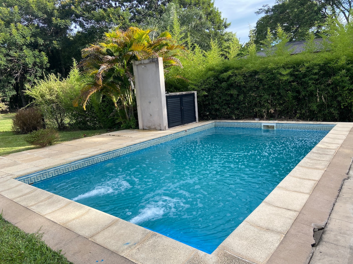 A private pool is set within a well-maintained outdoor area, featuring a clean, inviting water surface. Surrounding greenery includes lush plants and trees, providing shade. A small outdoor shower is present near the pool, enhancing the space for relaxation and enjoyment.
