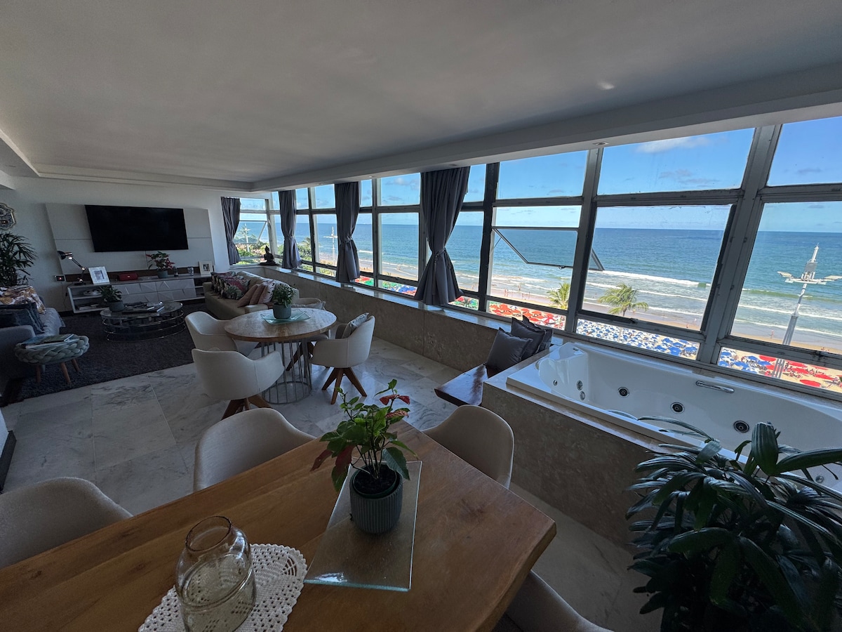 A bright living area features expansive windows that frame a stunning view of the ocean. A dining table, set for six, complements a cozy seating arrangement. In the corner, a jacuzzi awaits by the glass wall, inviting relaxation while overlooking the beach.