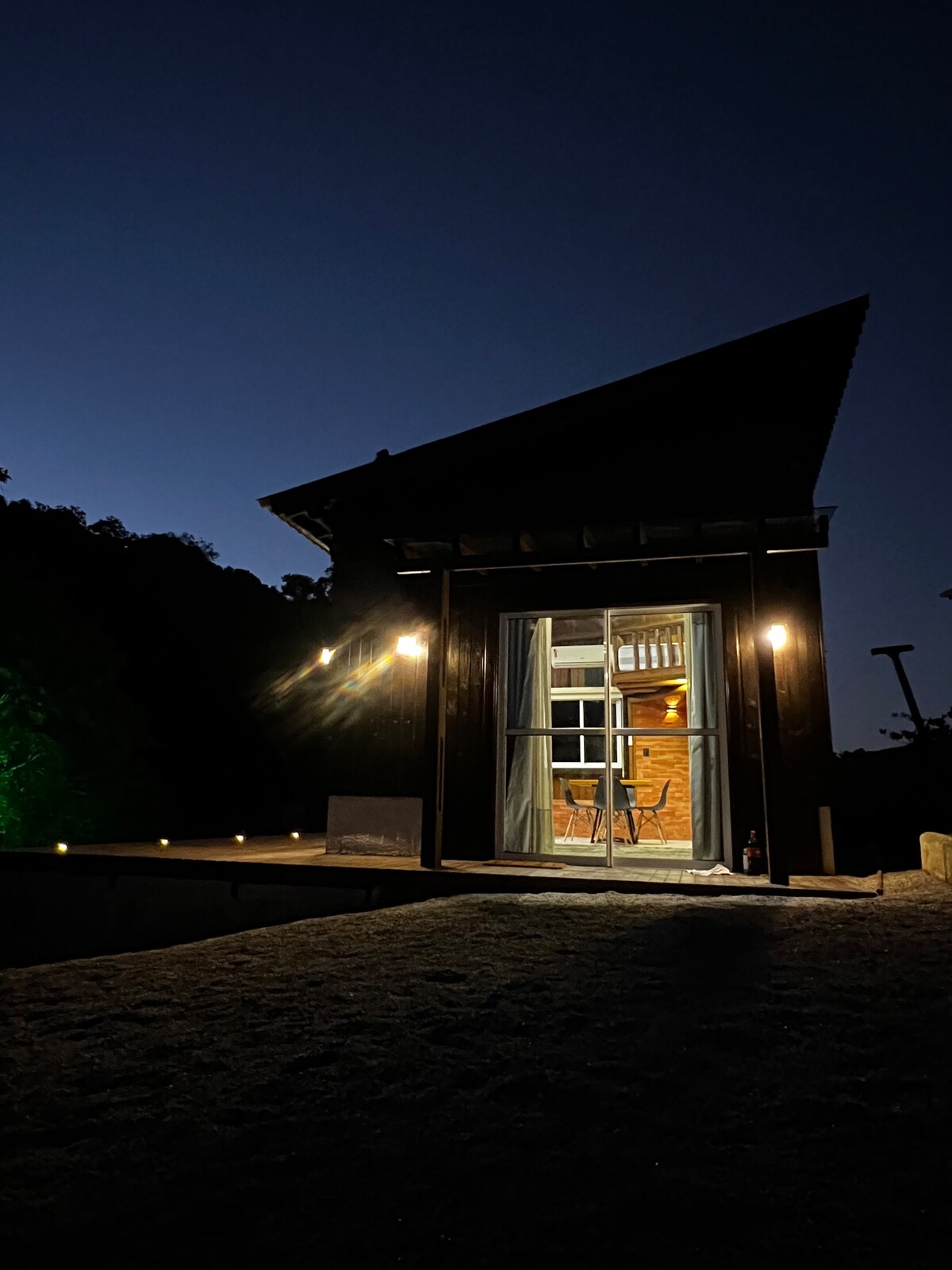 A unique cabin is illuminated by warm exterior lights, showcasing large glass doors that reveal a cozy interior. The structure features a distinctive peaked roof, framed by the surrounding dark landscape, blending architecture with nature under the evening sky.