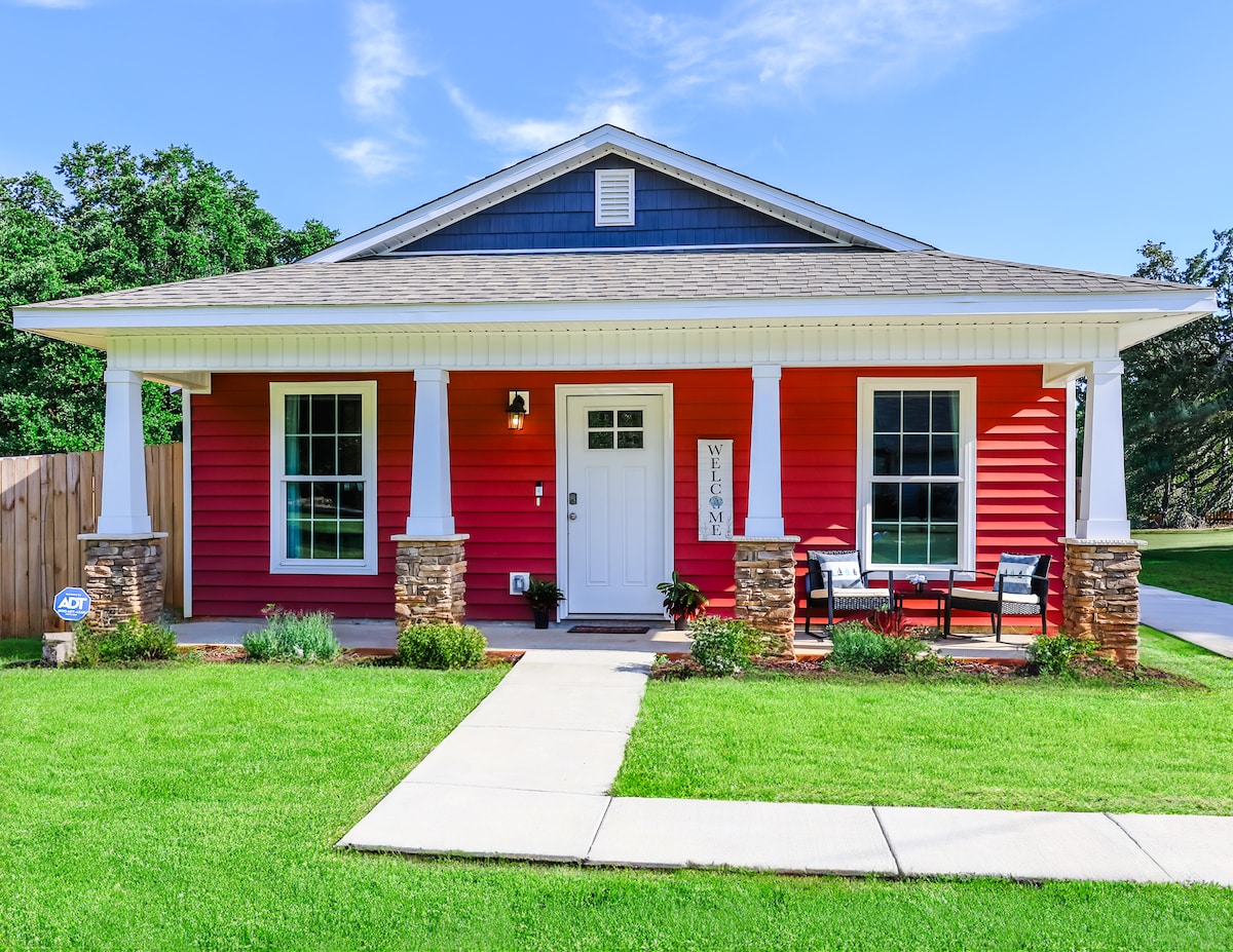 A freshly constructed home is shown with a red exterior and white trim. Two stone pillars frame the entrance, leading to a welcoming front door. A small seating area with chairs is visible on the porch, surrounded by a well-maintained lawn and landscaped garden.
