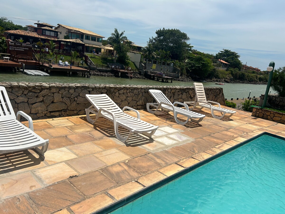 A stone patio surrounds a clear swimming pool, while several white lounge chairs are positioned for relaxation. The lush greenery and canal are visible in the background, creating a serene outdoor setting.
