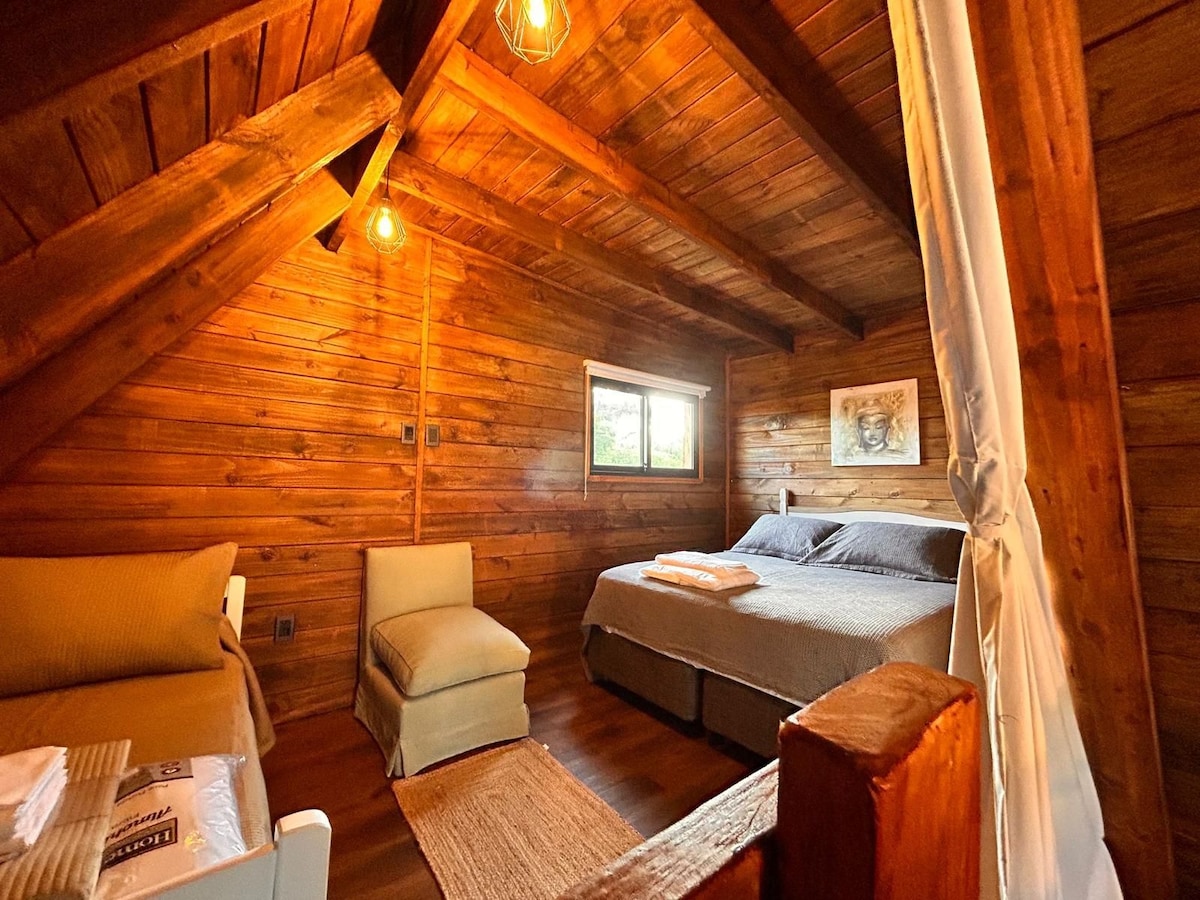 A cozy cabin interior features wooden walls and ceiling, with warm lighting from exposed bulbs. A spacious bed is positioned against one wall, complemented by a chair and a soft throw blanket. A window allows natural light to fill the room.