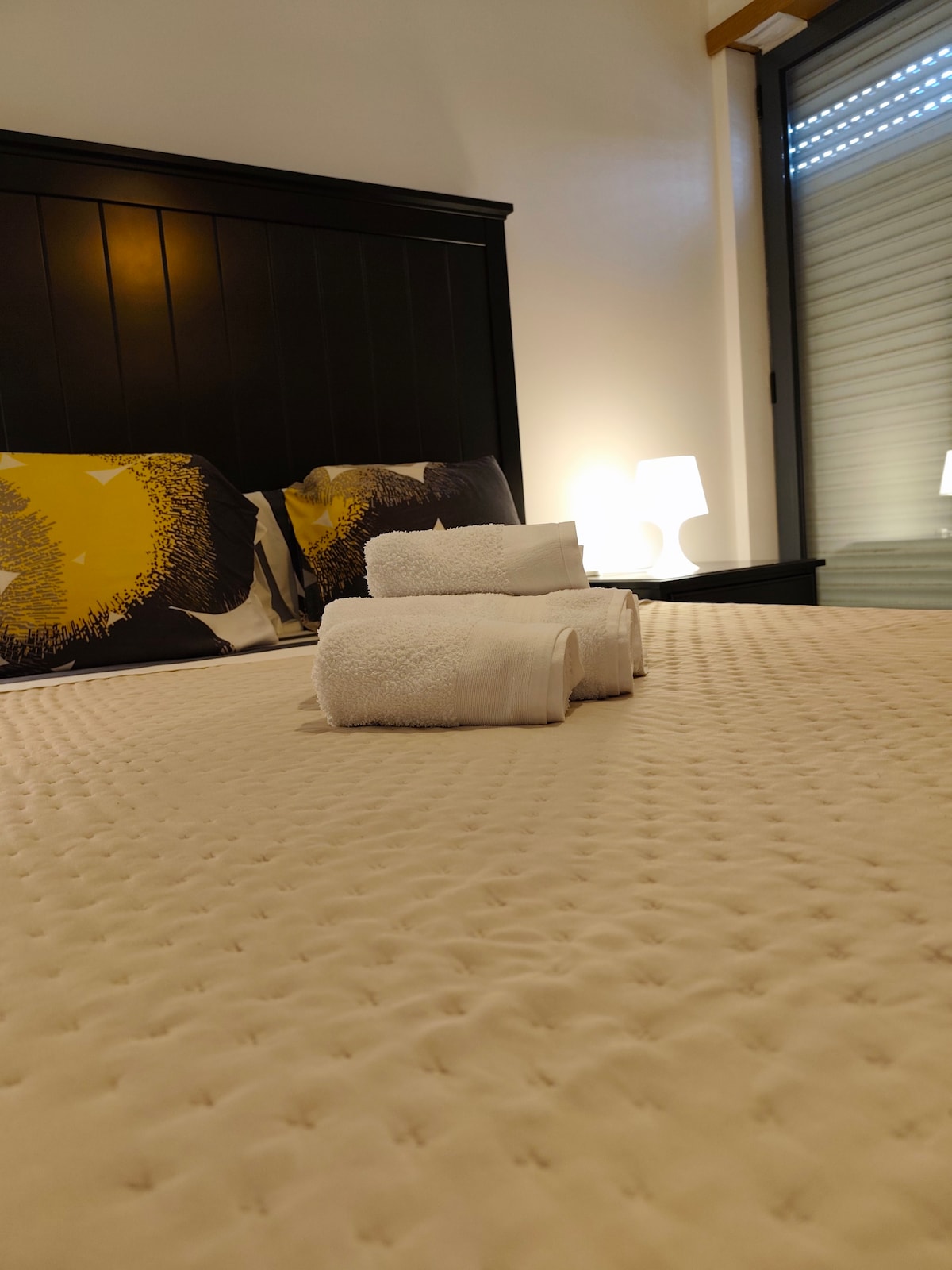 A comfortable bed is featured, adorned with a neutral-colored quilt. Two decorative pillows are positioned against the headboard. Nearby, a soft glow is provided by a table lamp, and fresh towels are neatly arranged on the bed, inviting a sense of relaxation.