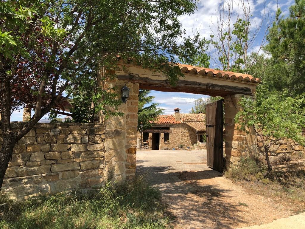 An inviting entrance is framed by rustic stone walls and a wooden gate, leading into a peaceful countryside retreat. Lush greenery surrounds the entryway, while a charming stone building is visible in the background under a clear sky.
