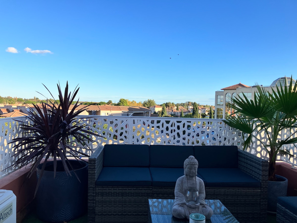 A spacious balcony is featured, adorned with a comfortable seating area surrounded by potted plants. A decorative statue sits on a low table, offering a serene contrast to the clear blue sky and distant rooftops. The intricate balcony railing provides a unique design element.