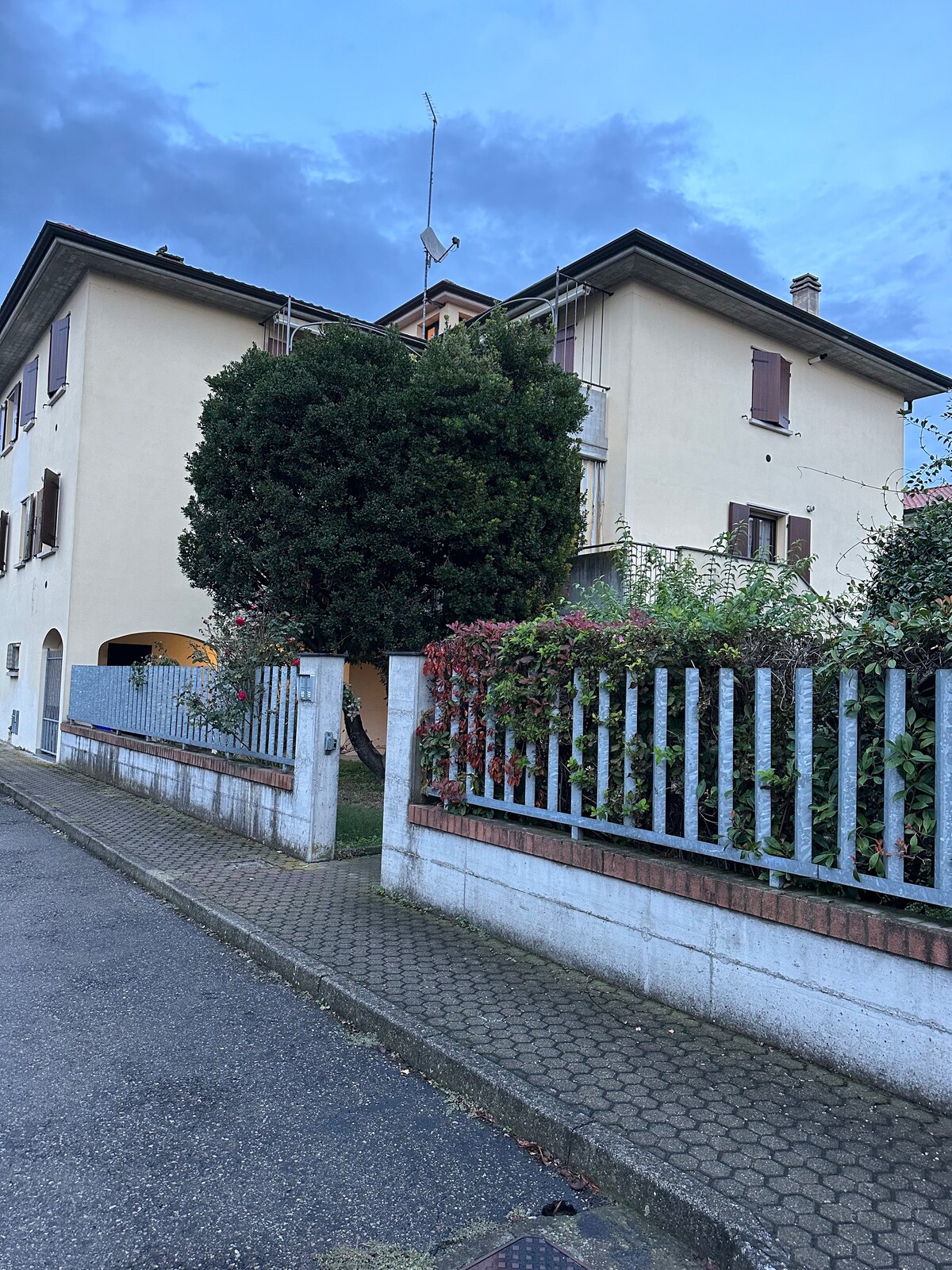 A modern building features a light-colored facade and multiple windows adorned with brown shutters. A lush green shrub separates the entrance from a cobblestone path, while a gated entry provides access. Surrounding plants add greenery to the scene, with a cloudy sky above.