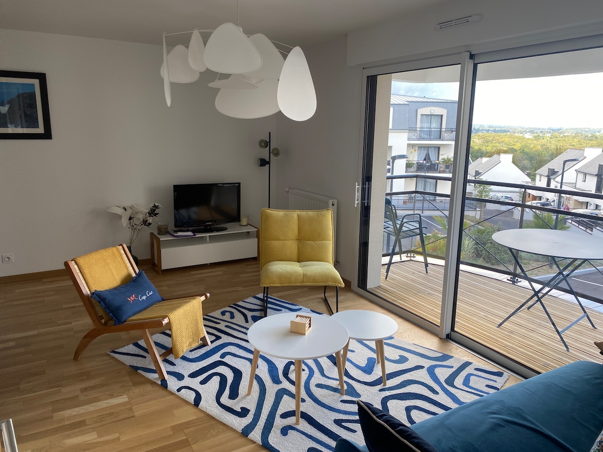 A bright living area features modern furnishings, including a yellow armchair and a wooden lounge chair. A flat-screen television is positioned on a white console. Large glass doors open to a balcony with outdoor seating, allowing natural light to enhance the atmosphere. A patterned area rug lies on the wooden floor.