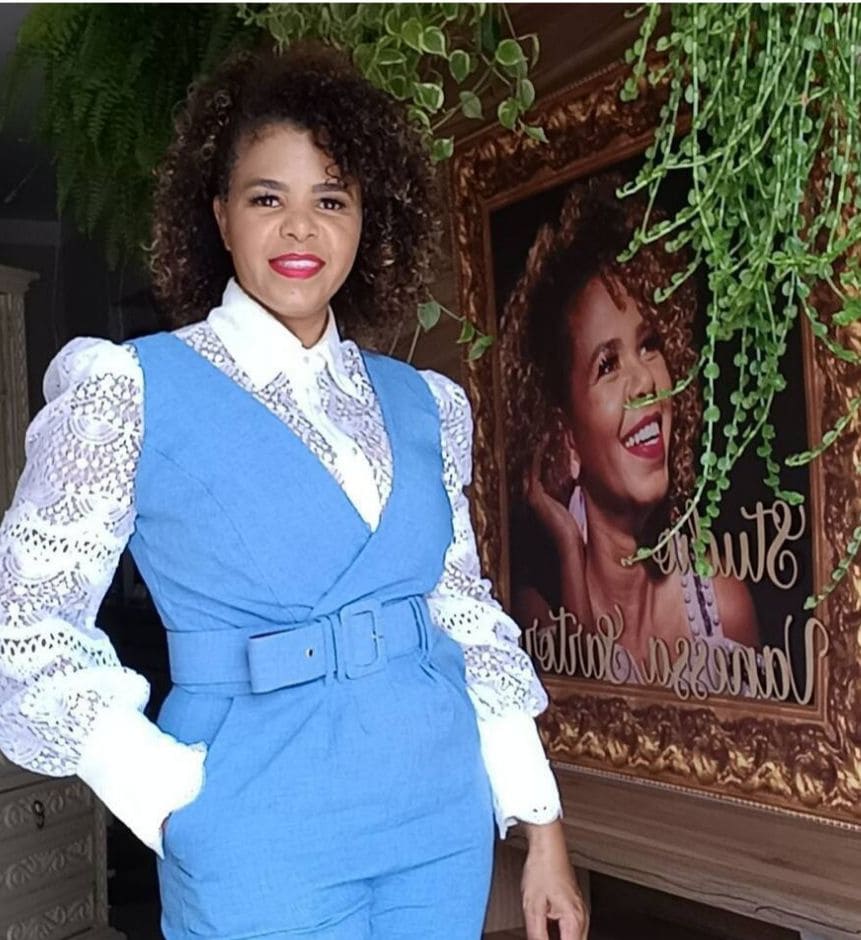 A stylish outfit featuring a blue suit with a belted waist is complemented by long, lace-trimmed white sleeves. In the background, a large portrait and decorative greenery add visual interest to the setting.