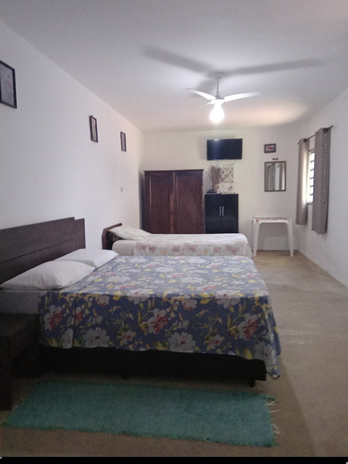 A simple sleeping area is presented, featuring a spacious bed with a floral-patterned blanket. A second bed is positioned alongside it, and light is provided by a ceiling fan above. A wooden cabinet and a small side table are visible, enhancing the functionality of the space.
