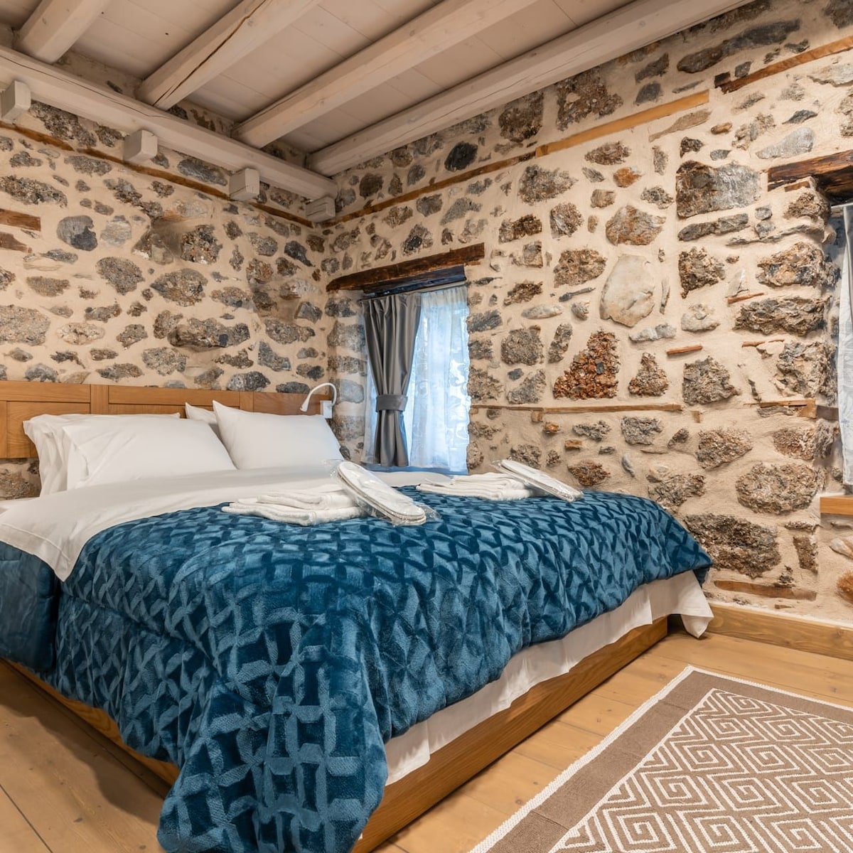 A cozy sleeping area is surrounded by rustic stone walls, showcasing a spacious bed with a textured blue blanket. Two plush pillows are arranged at the head of the bed, and soft white towels are neatly folded nearby. Natural light filters through a window dressed with light curtains.