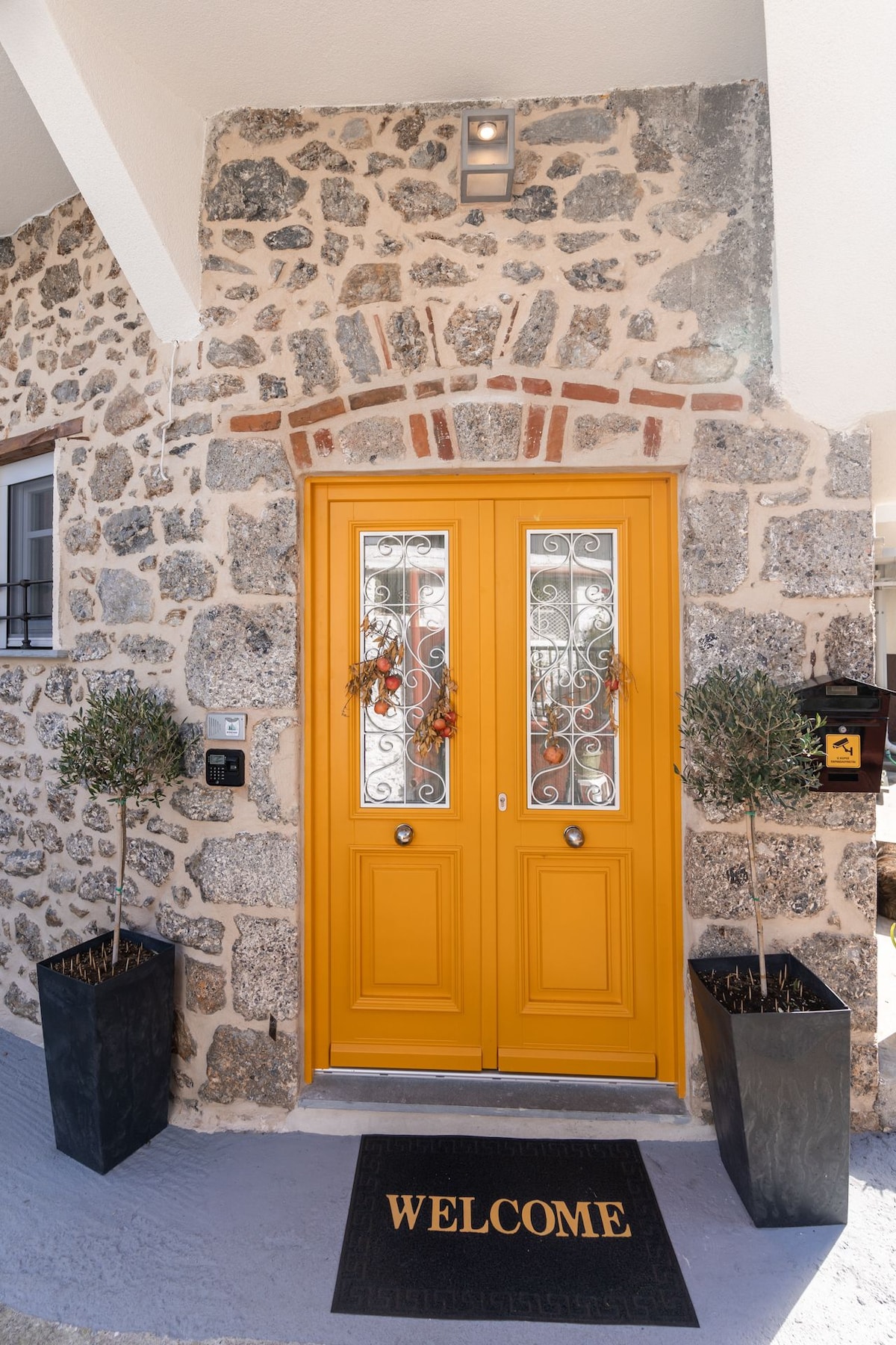 A charming stone exterior features a pair of bright yellow doors with decorative glass panels. On either side, tall planters containing olive trees add a touch of greenery. A welcome mat positioned at the entrance invites guests into the home.