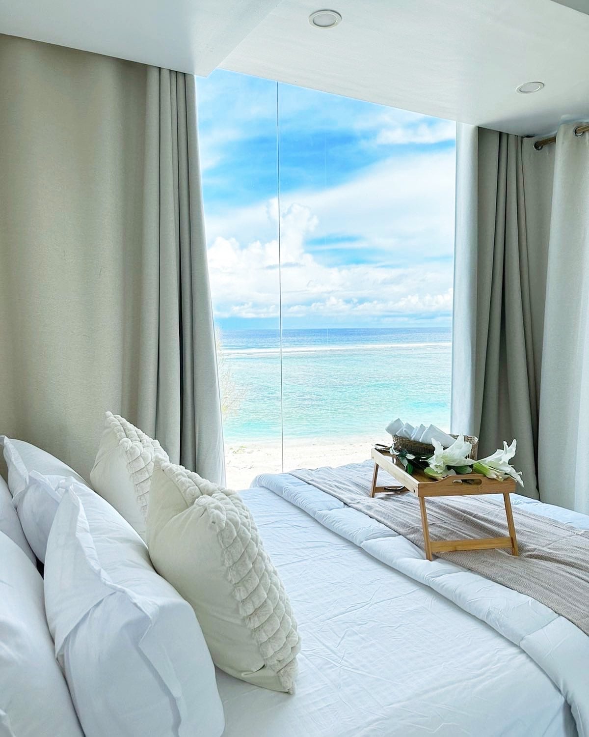 A spacious bedroom features a large bed adorned with decorative pillows. A tray with fresh flowers is placed gently on the bed. Floor-to-ceiling windows provide a stunning view of the beach and ocean, filling the room with natural light.