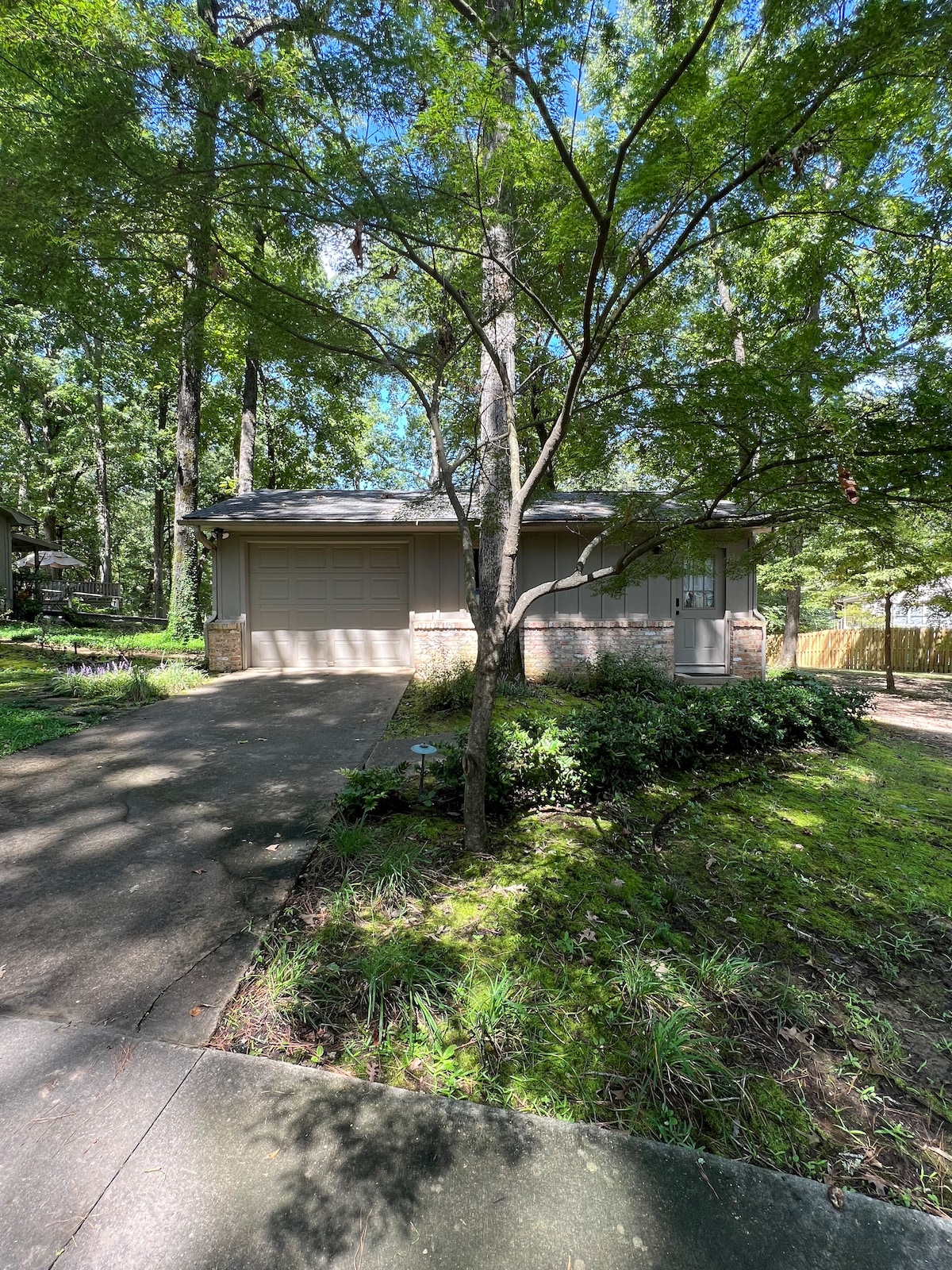 A serene exterior of a detached guest home is visible, set among lush green trees. A paved driveway leads to a two-car garage, surrounded by natural landscaping and a grassy area. The structure features soft earth tones, blending harmoniously with the wooded surroundings.
