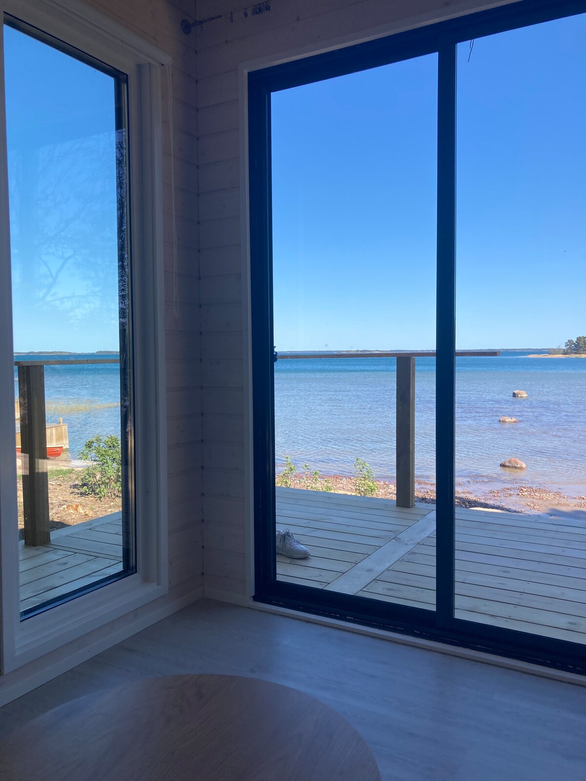 Large windows frame a serene view of the water and rocky shoreline, allowing natural light to fill the space. A wooden deck is visible just outside, creating a seamless transition between indoor and outdoor areas.