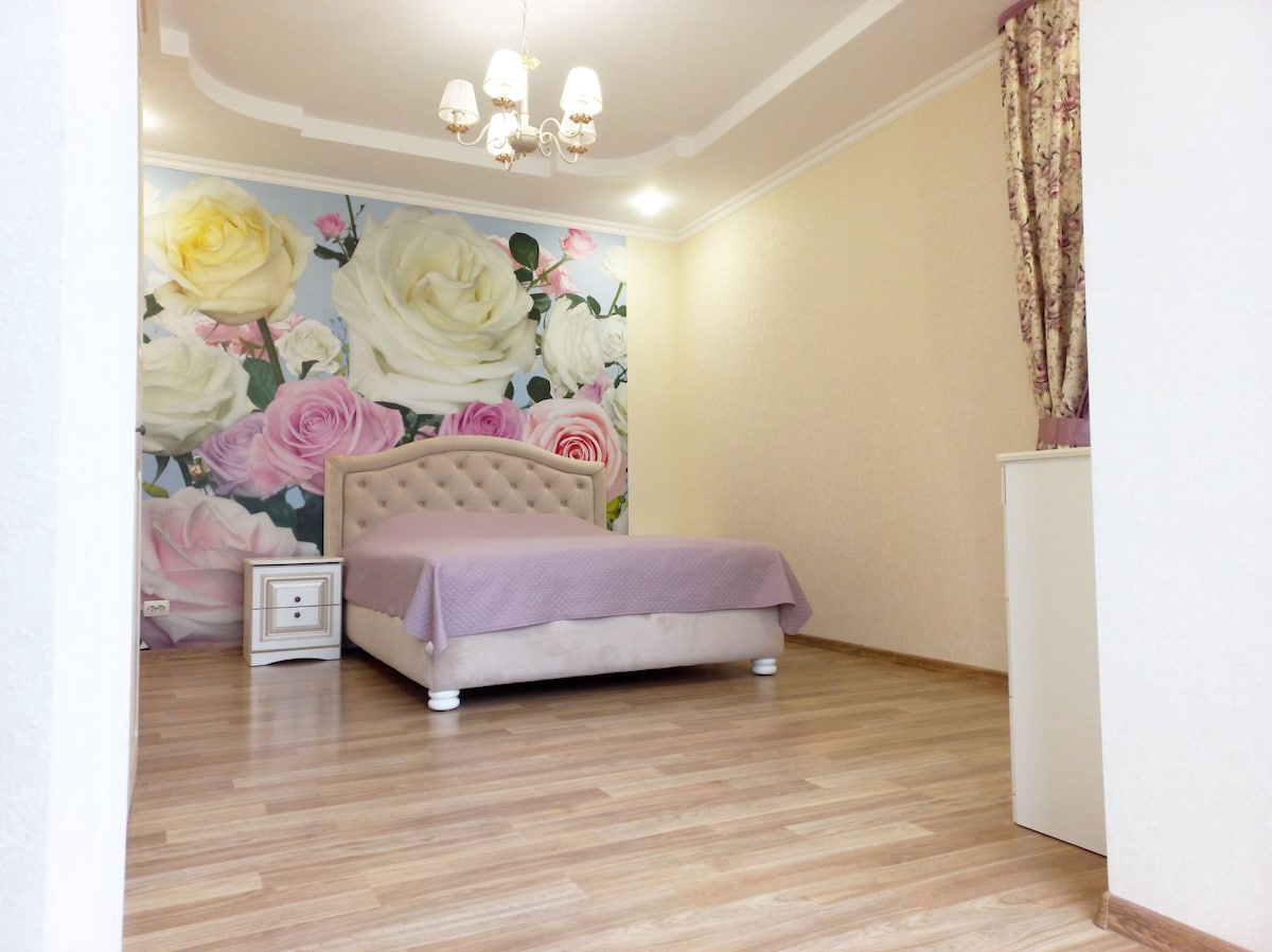 A spacious bedroom features a light-colored bed with a tufted headboard, complemented by a purple bedspread. A floral wallpaper of roses adorns one wall, creating a soft ambiance. Natural light is provided by a window with floral curtains.