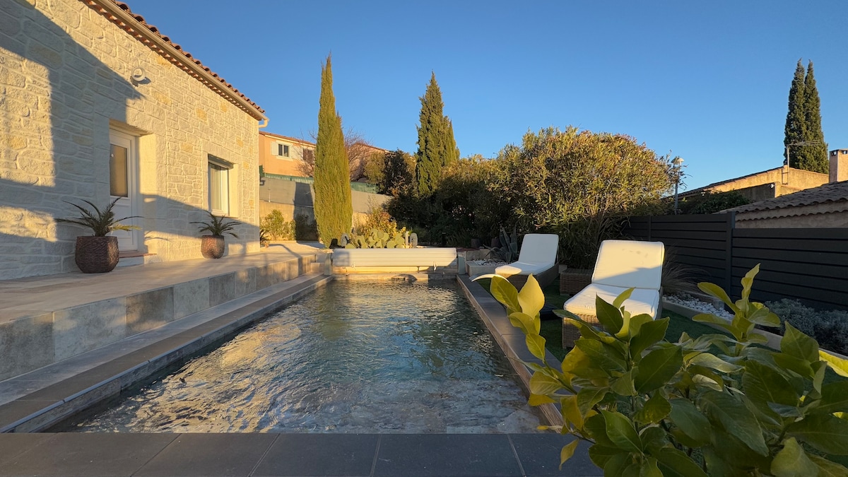 An outdoor area features a luxurious swimming pool with a smooth stone surround, framed by landscaping and tall trees. Two lounge chairs are positioned beside the pool, providing a tranquil spot for relaxation under the clear blue sky.
