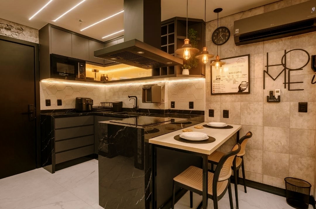 A modern kitchen features sleek cabinetry with dark finishes and integrated lighting. A central island is surrounded by two wicker chairs. Countertop appliances and kitchen essentials are organized for convenience. Warm tones from pendant lights enhance the stylish ambiance, while air conditioning ensures comfort.