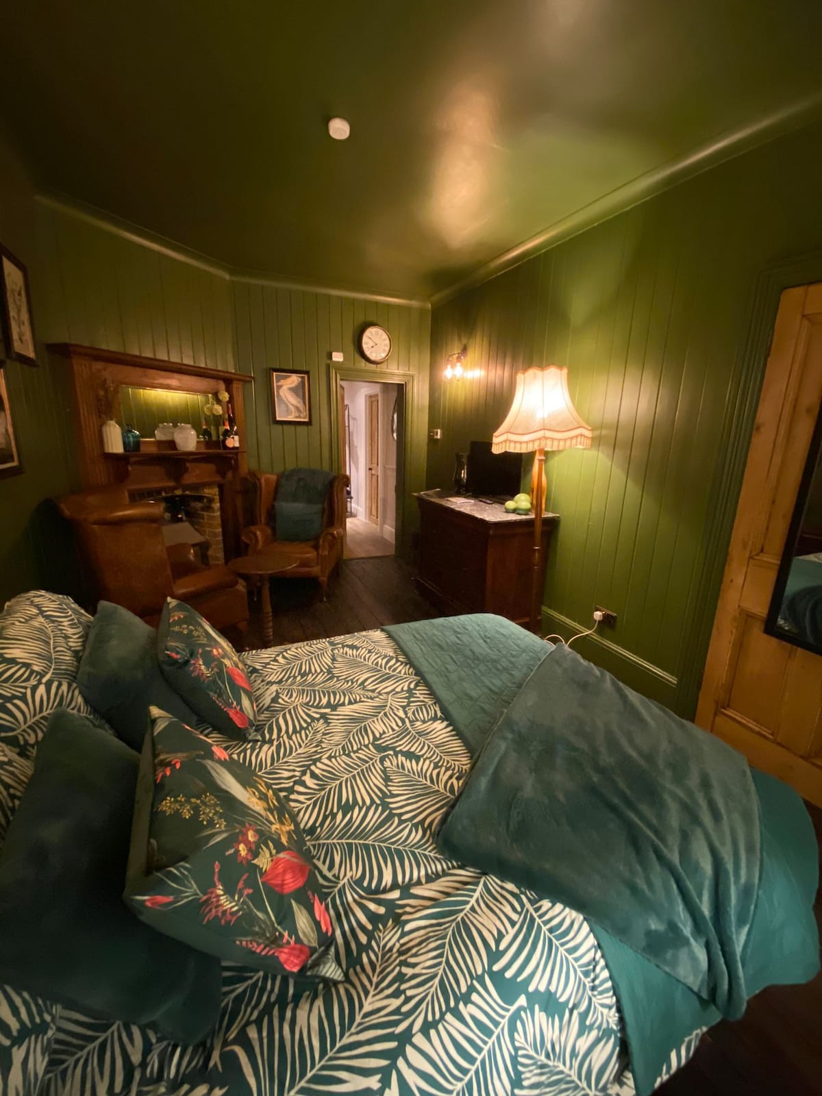 A cozy bedroom features deep green walls and a stylish wooden dresser. A spacious bed is adorned with tropical-patterned pillows and a soft coverlet. A vintage lamp and mirror enhance the inviting ambiance. In the background, a doorway leads to the en-suite shower room.